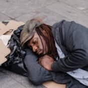 A homeless man sleeping on the street with cardboard signs and belongings.